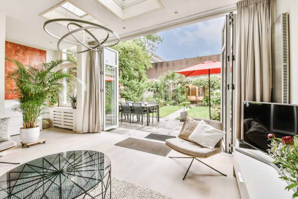 Bright modern living room opening onto a lush backyard with a dining patio, red umbrellas, green lawn, and vibrant landscaping under a blue sky.