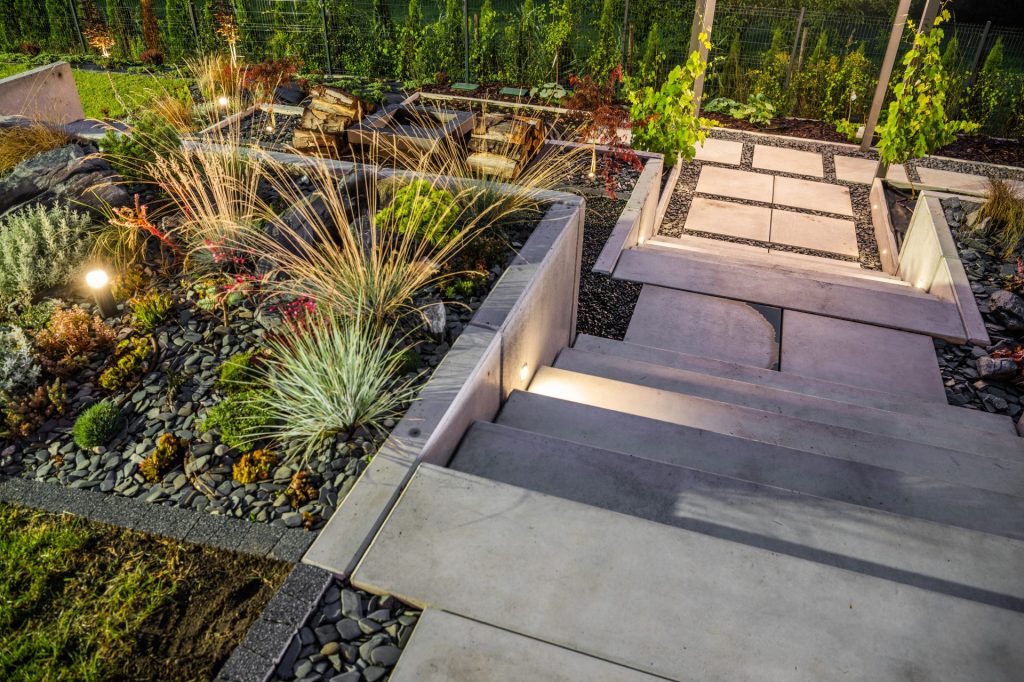 Modern backyard garden with concrete steps, ambient lighting, and landscaped rock beds featuring ornamental grasses and colorful plants.