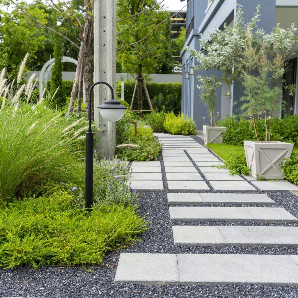 Modern garden walkway with rectangular concrete pavers set in gravel, surrounded by ornamental grasses, shrubs, and potted plants beside a contemporary home.
