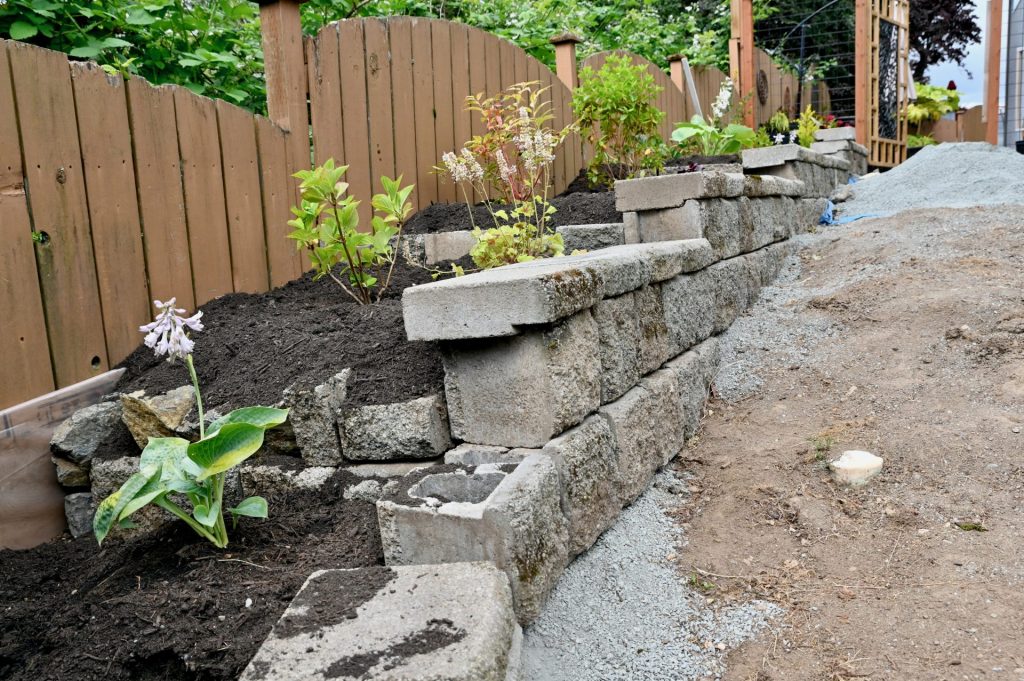 Tiered retaining wall built with concrete blocks, partially under construction, with newly planted shrubs and flowers in fresh soil along a wooden fence.