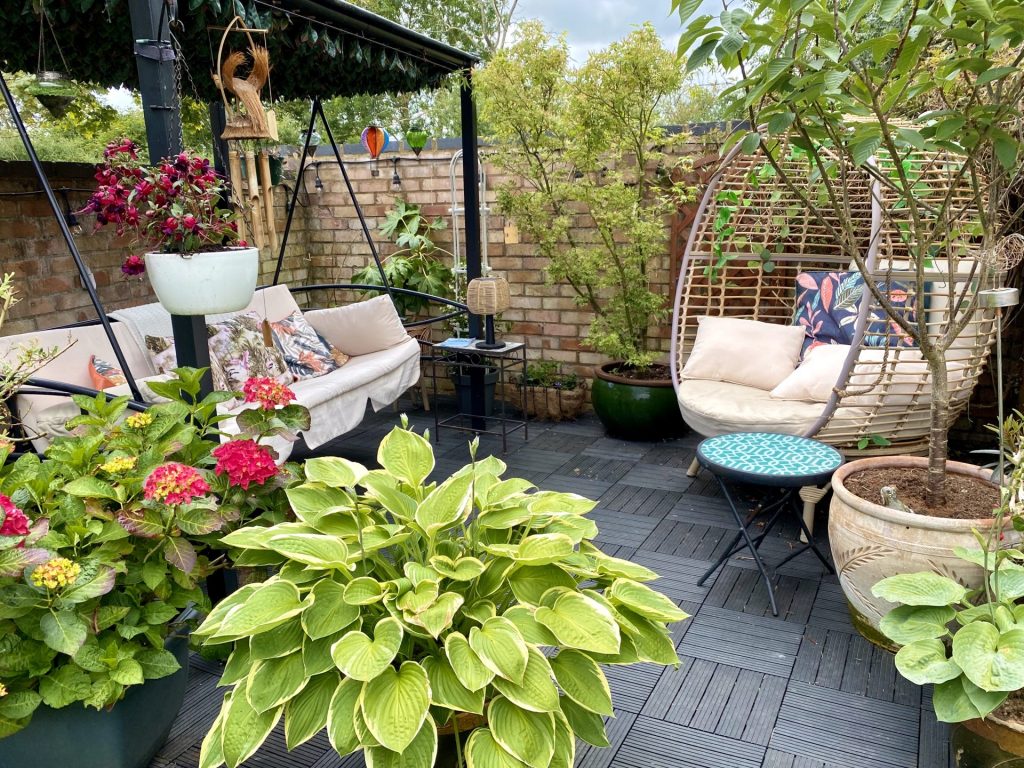 Cozy backyard patio with cushioned seating, lush container plants, and a hanging swing chair, surrounded by a brick wall and garden greenery.