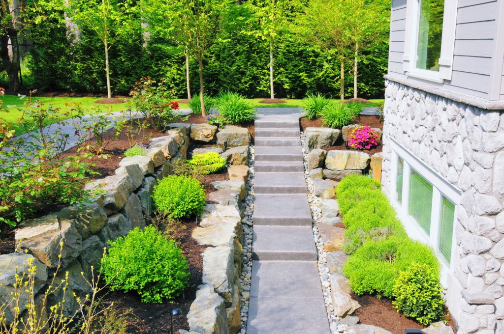 Stone steps flanked by garden beds with neatly trimmed shrubs, flowering plants, and large rocks, leading to a lush lawn bordered by trees in a well-landscaped yard beside a stone-clad home
