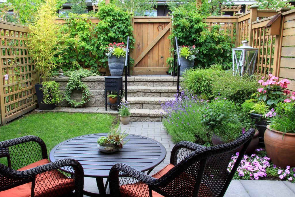 Charming fenced backyard with lush greenery, colorful flowers, potted plants, and a bistro table set on a stone patio. Stone steps lead to a wooden gate framed by climbing vines.