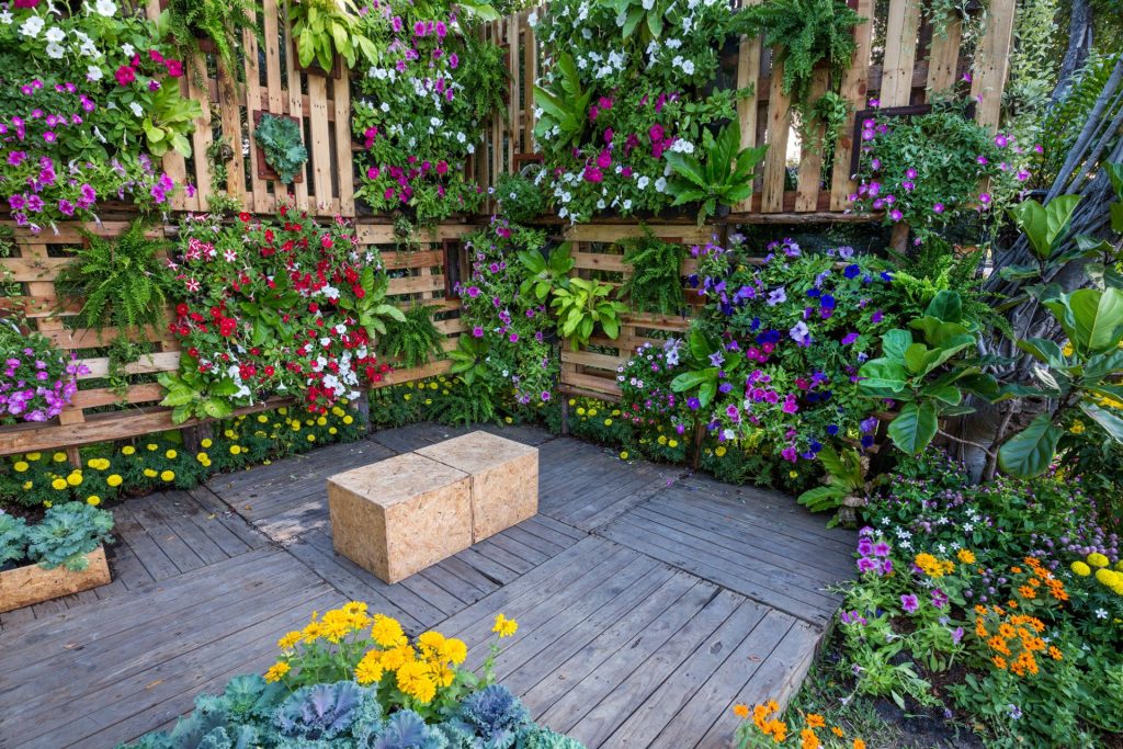 Colorful vertical garden made from wooden pallets filled with vibrant flowers and lush greenery, surrounding a wooden deck with simple box-style seating and more blooms planted along the floor.