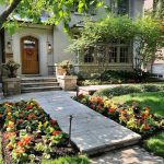 Elegant front yard garden with a stone walkway leading to a wooden door, lined with vibrant red and yellow flowers, neatly trimmed grass, and shaded by mature trees and shrubs.