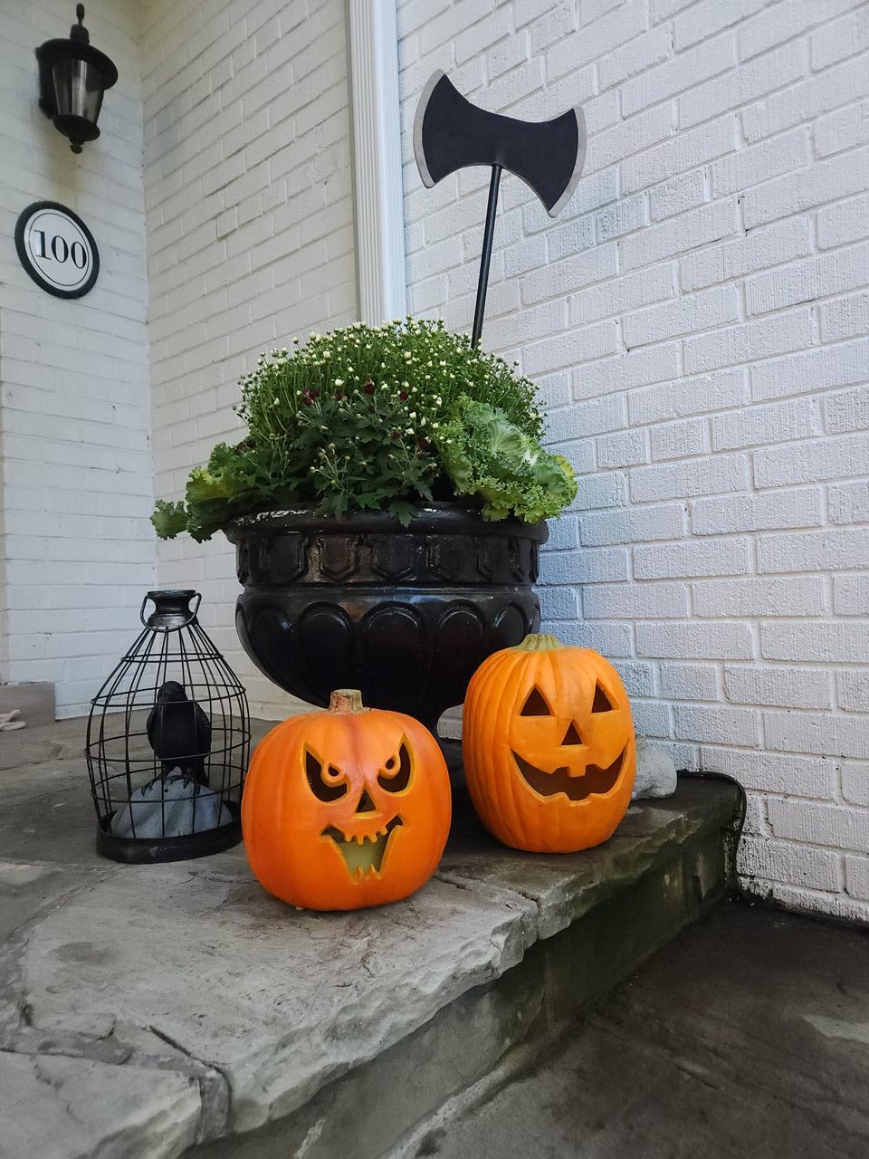 Two carved pumpkins on porch with decor and plants.
